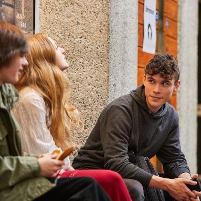 A boy wearing a grey jumper sits and faces a girl in a white shirt and a boy in a green jacket. He has a smile on his face.